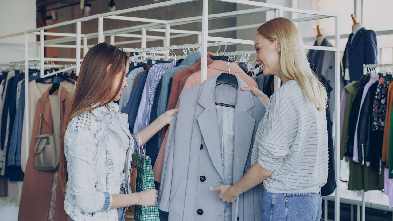 Two women shopping in a clothing store, examining a stylish gray blazer on a rack.